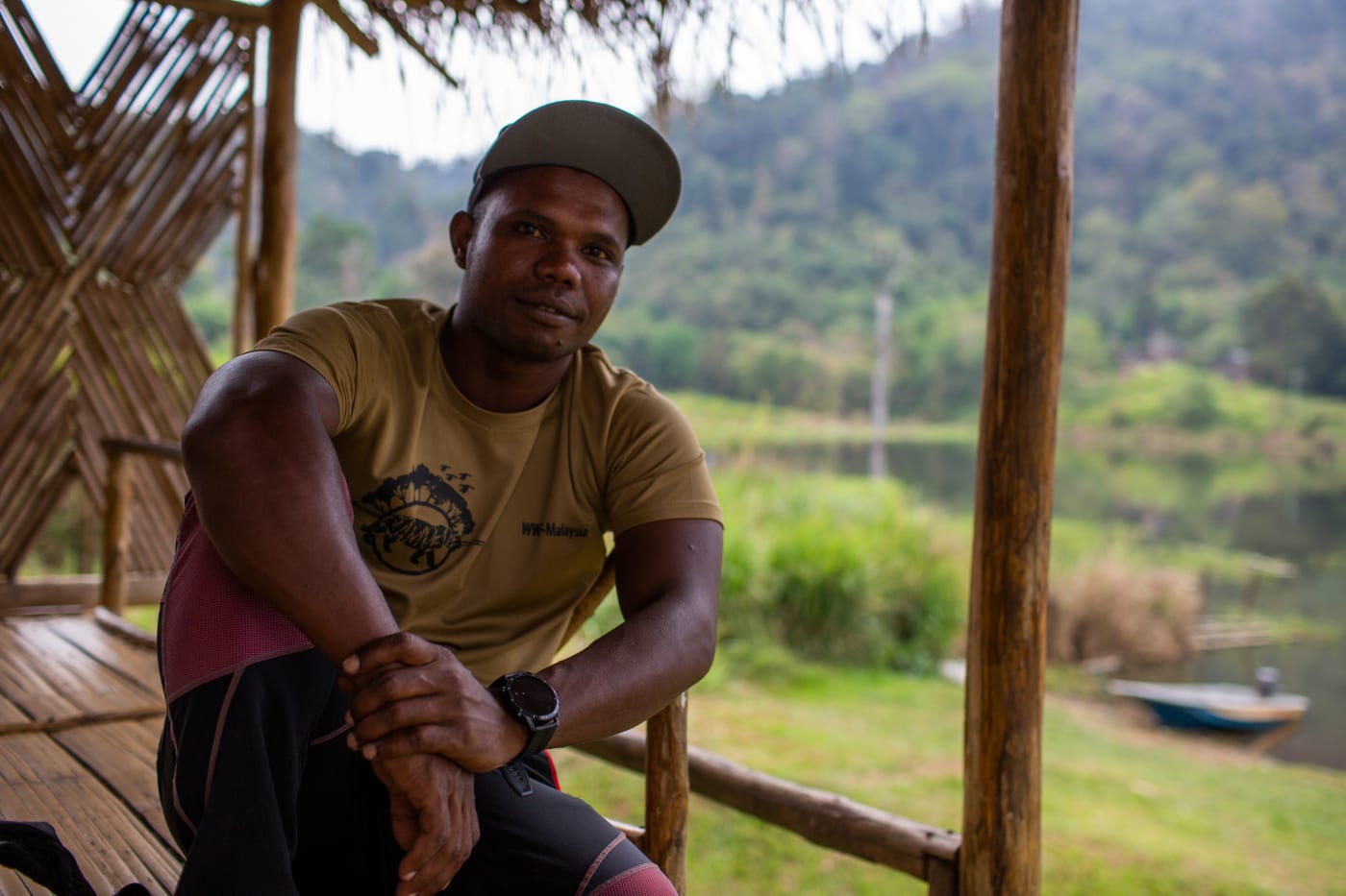 Merapi sits on a wooden deck looking into the camera. He is wearing a yellow short and cap. Behind him are forest-covered mountains.