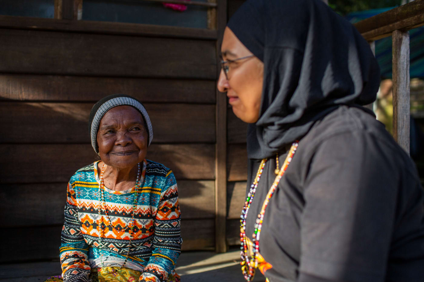 Two women sit inside a room with wood panelled walls. They are in conversation and smiling. Both are wearing colourful beaded necklaces.