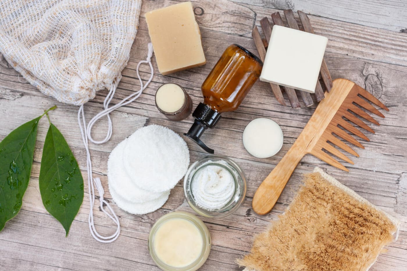 A selection of reusable bathroom accessories lay on a wooden surface. Included are bars of soap, a wooden comb, cloth make-up wipes and a glass bottle of lotion.