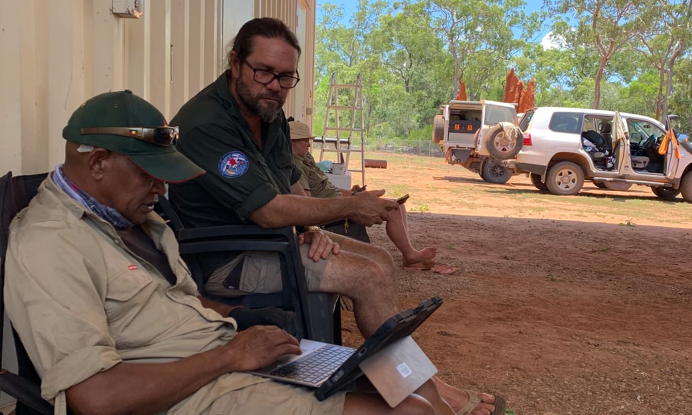 Wuthathi Ranger Clayton Enoch sits with another Ranger, who is using a laptop. They are sitting undercover but outside. The dirt is a red colour. Trees, blue sky and a four-wheel-drive car can be seen in the background.