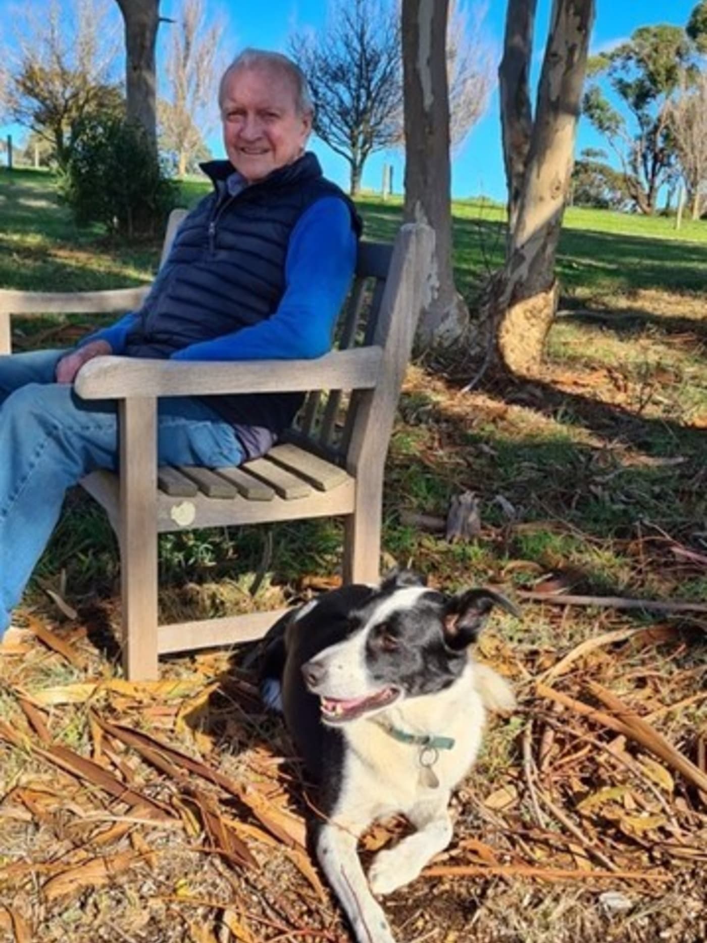 Tony sits in a park on a wooden bench, with a black and white dog laying near his feet. He is smiling and wearing jeans, a blue top and a black puffer vest.