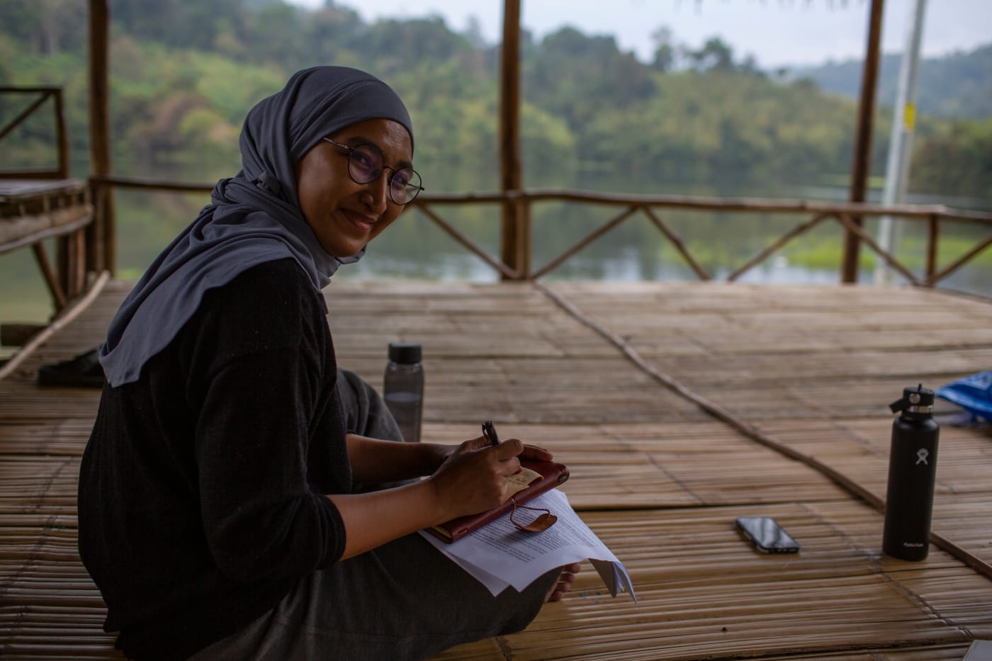 Senior Community Engagement and Education Officer Umi sits on a wooden deck, writing on a notebook. She has turned to face the camera and is smiling.