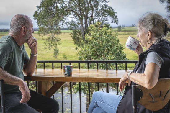 A man and a woman sit on their wooden deck overlooking green paddocks and trees. The man is rubbing his chin in thought and the woman is raising a mug of tea to her lips.