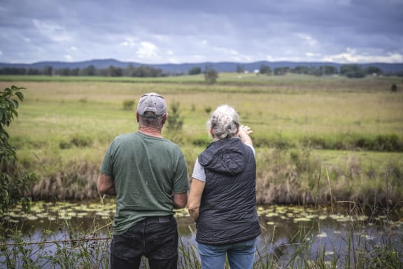 Couple Lyn Thomson and Steve Posselt have their backs to the camera as they look out over the fields and trees of their property. In front of them is a pond with lilypads, and dark rain clouds loom overhead in the background.