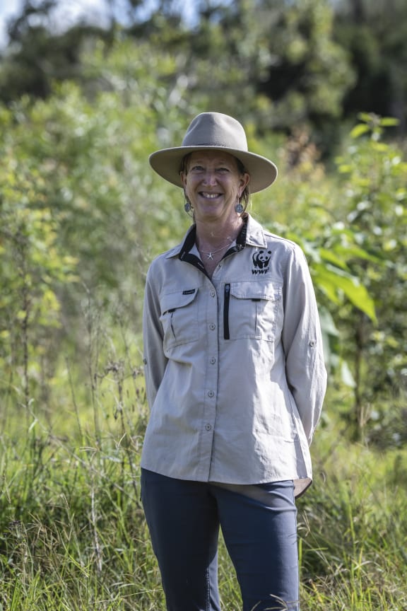 A woman smiles at the camera. She is wearing navy pants, a wide-brimmed hat and a taupe shirt with a WWF panda logo on it. Behind her are native trees.