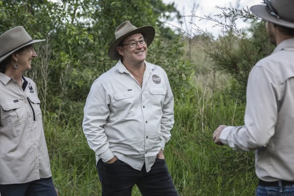 Three people laugh and talk together amongst green trees and knee-high grass. They are all wearing wide-brimmed hats to protect themselves from the sun.