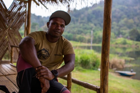 Merapi sits on a wooden deck looking into the camera. He is wearing a yellow short and cap. Behind him are forest-covered mountains.