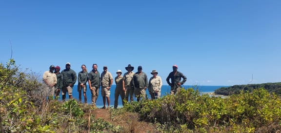 A group of Wuthathi Rangers stand atop a sand dune facing the camera. They are wearing ranger clothing. They are surrounded by short green vegetation, and the ocean and blue sky is in the background.